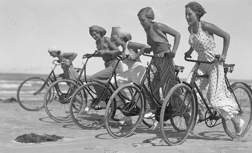 Fem piger i sommertøj cykler langs stranden.